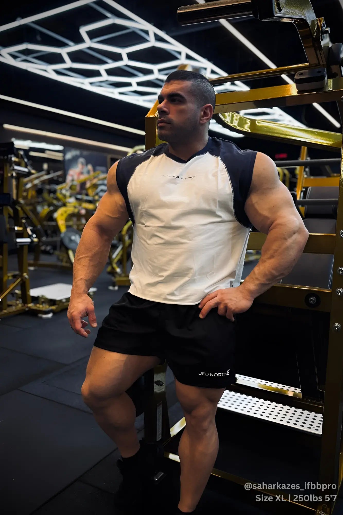 A muscular man wearing a white and navy raglan sleeveless tank top with gold-toned gym equipment in the background