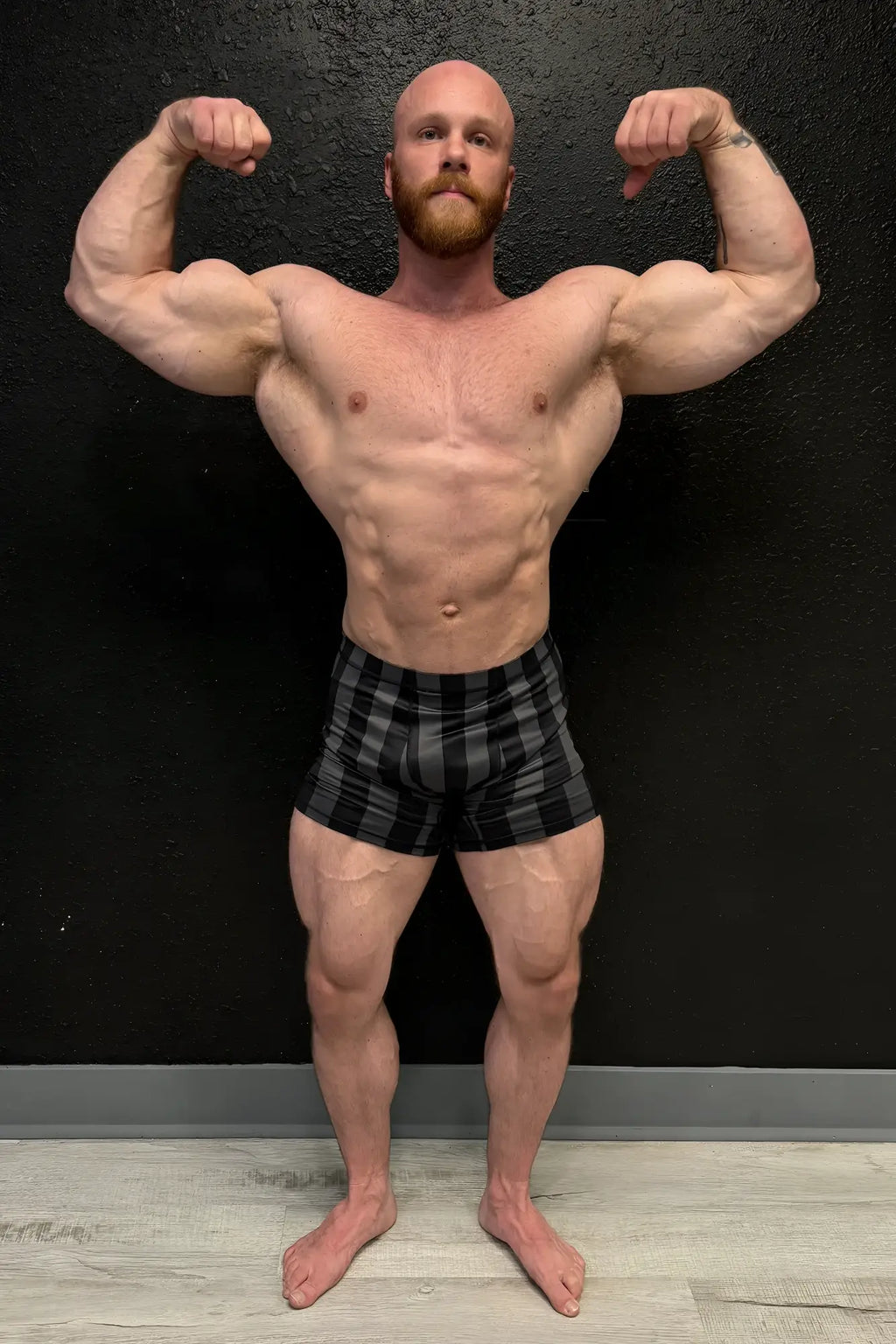 A muscular man with a bald head and red beard flexes wearing black and gray plaid retro bodybuilding shorts