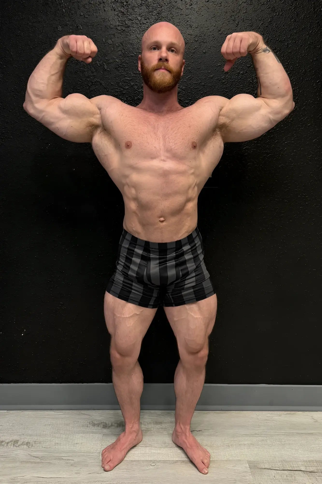 A muscular man with a bald head and red beard flexes wearing black and gray plaid retro bodybuilding shorts