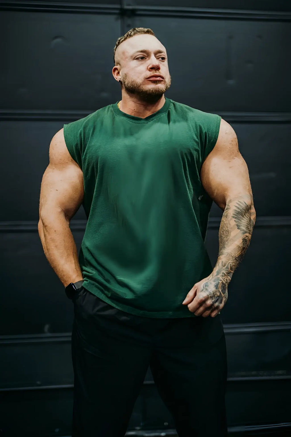 A man wearing a dark green retro oversized muscle tee sleeveless athletic shirt paired with black pants