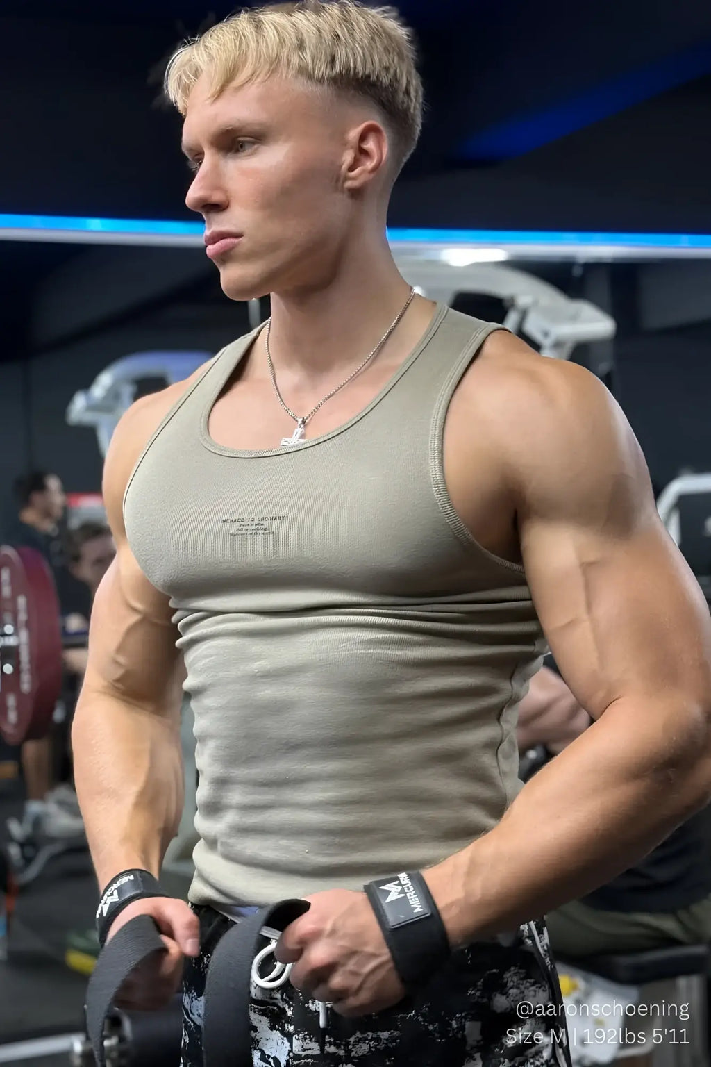 A muscular man wearing a ribbed khaki muscle tank top with subtle chest text and black weightlifting wrist wraps