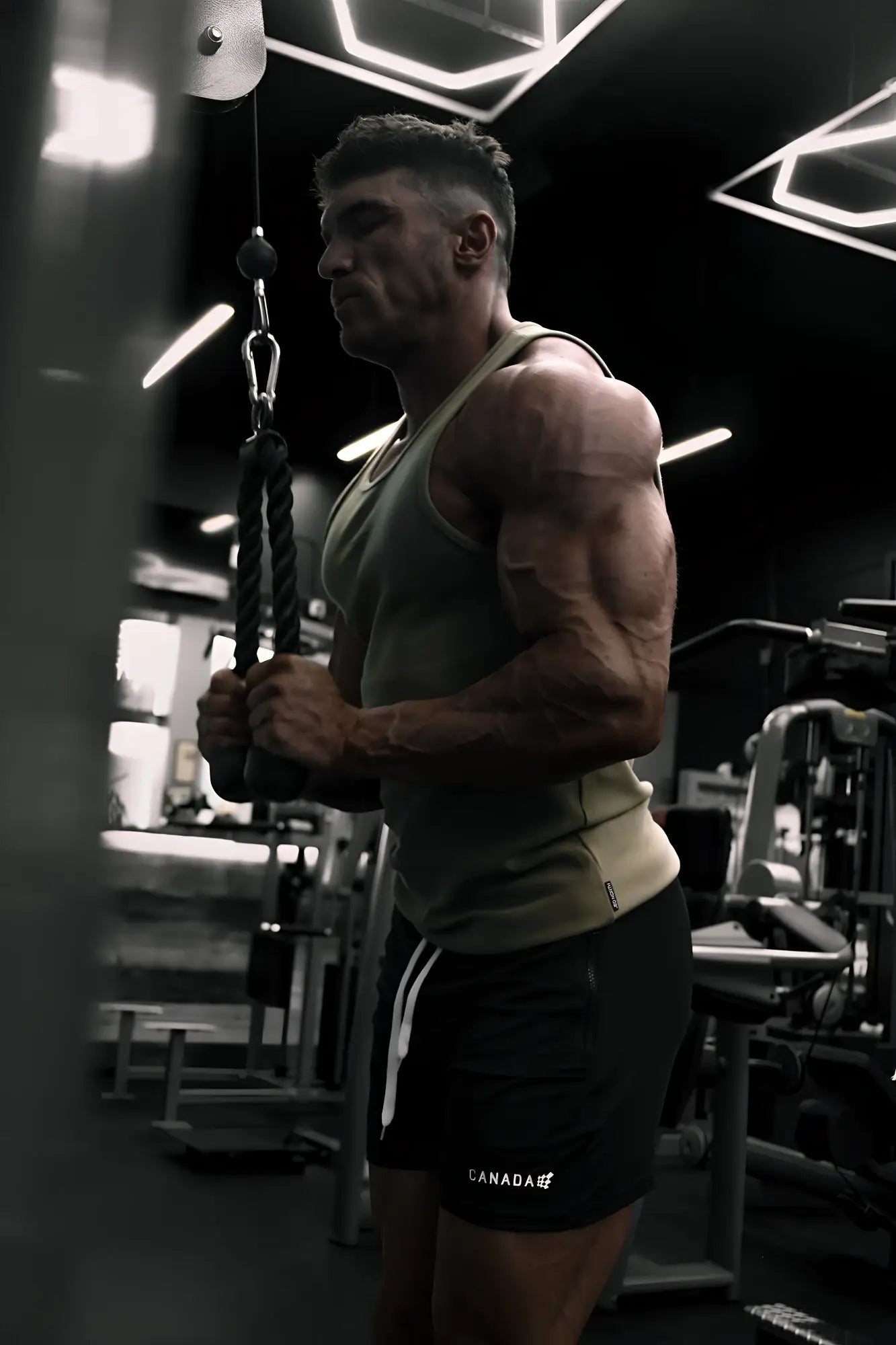 A muscular man wearing a khaki ribbed muscle tank top holding gym ropes during a workout session