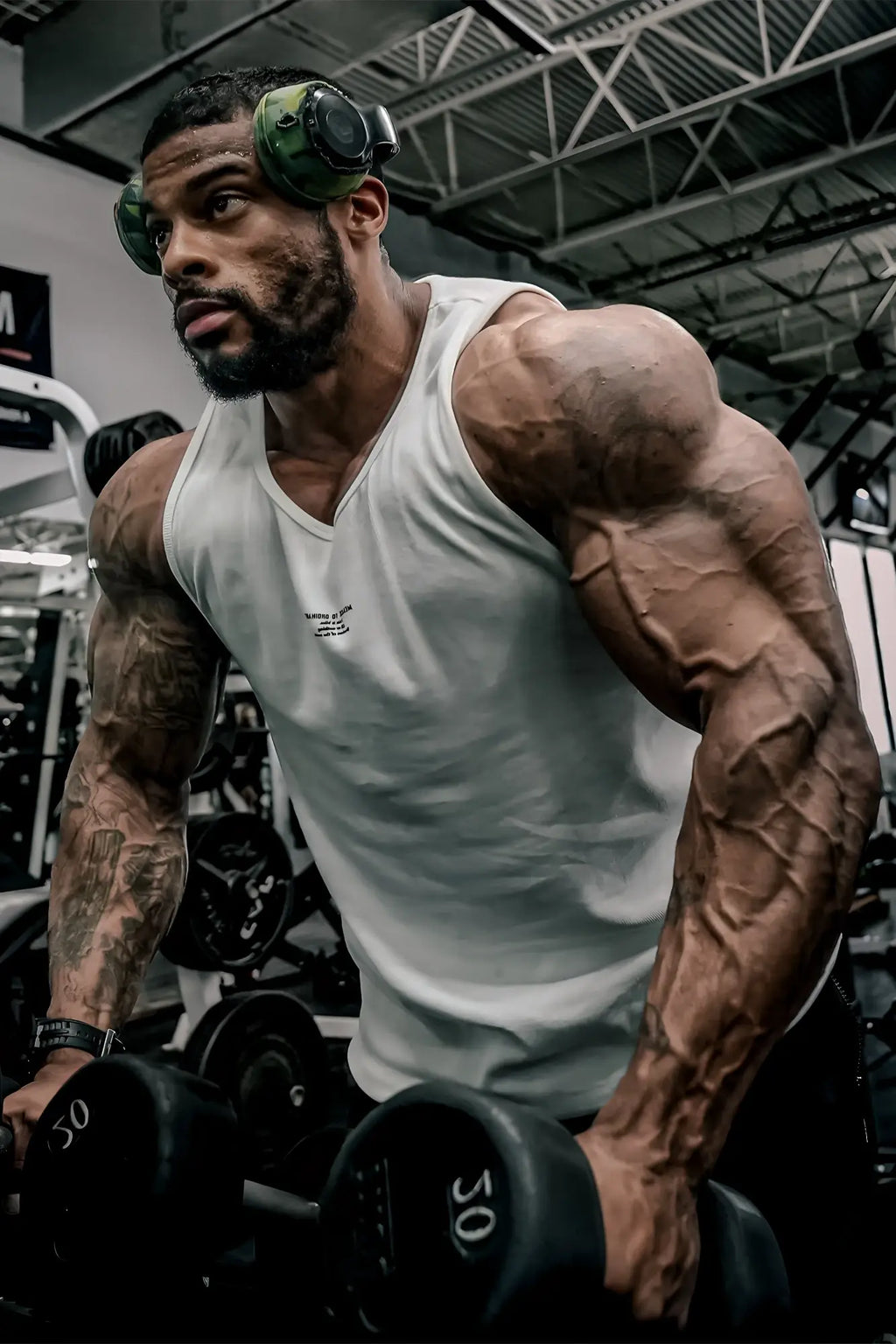 A muscular man wearing a white ribbed muscle tank top and green headphones holding dumbbells in a gym