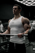 A muscular man wearing a white ribbed muscle tank top holding a barbell in a gym workout setting