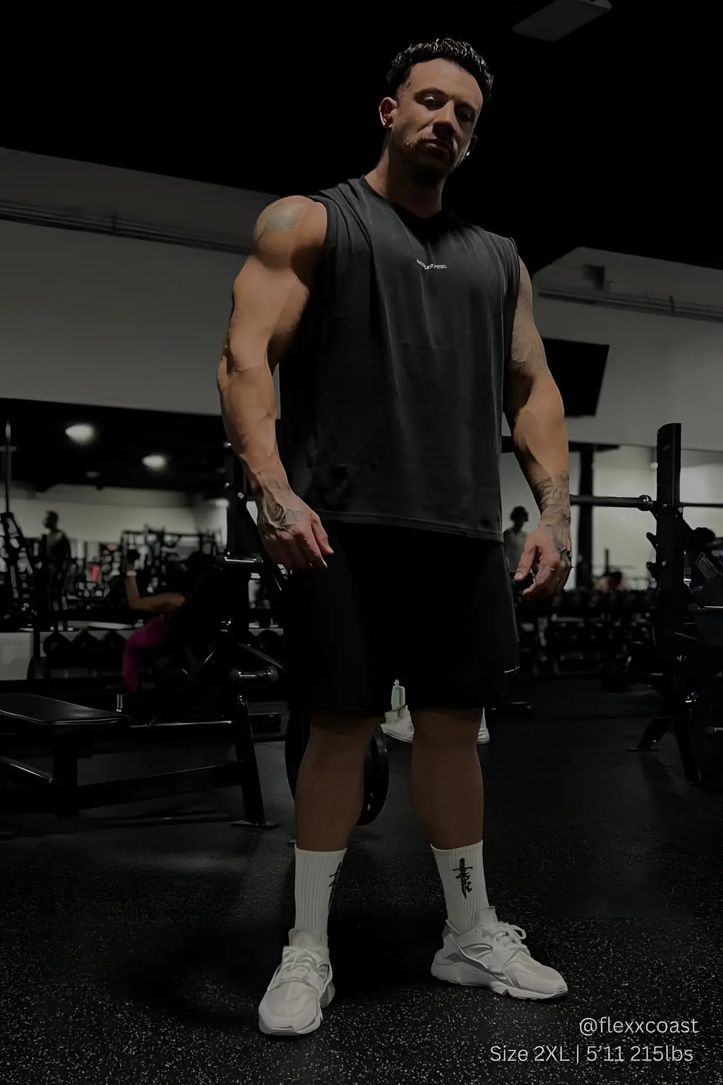 A muscular man wearing a black jed north sleeveless box cut tank top with black shorts and white athletic shoes