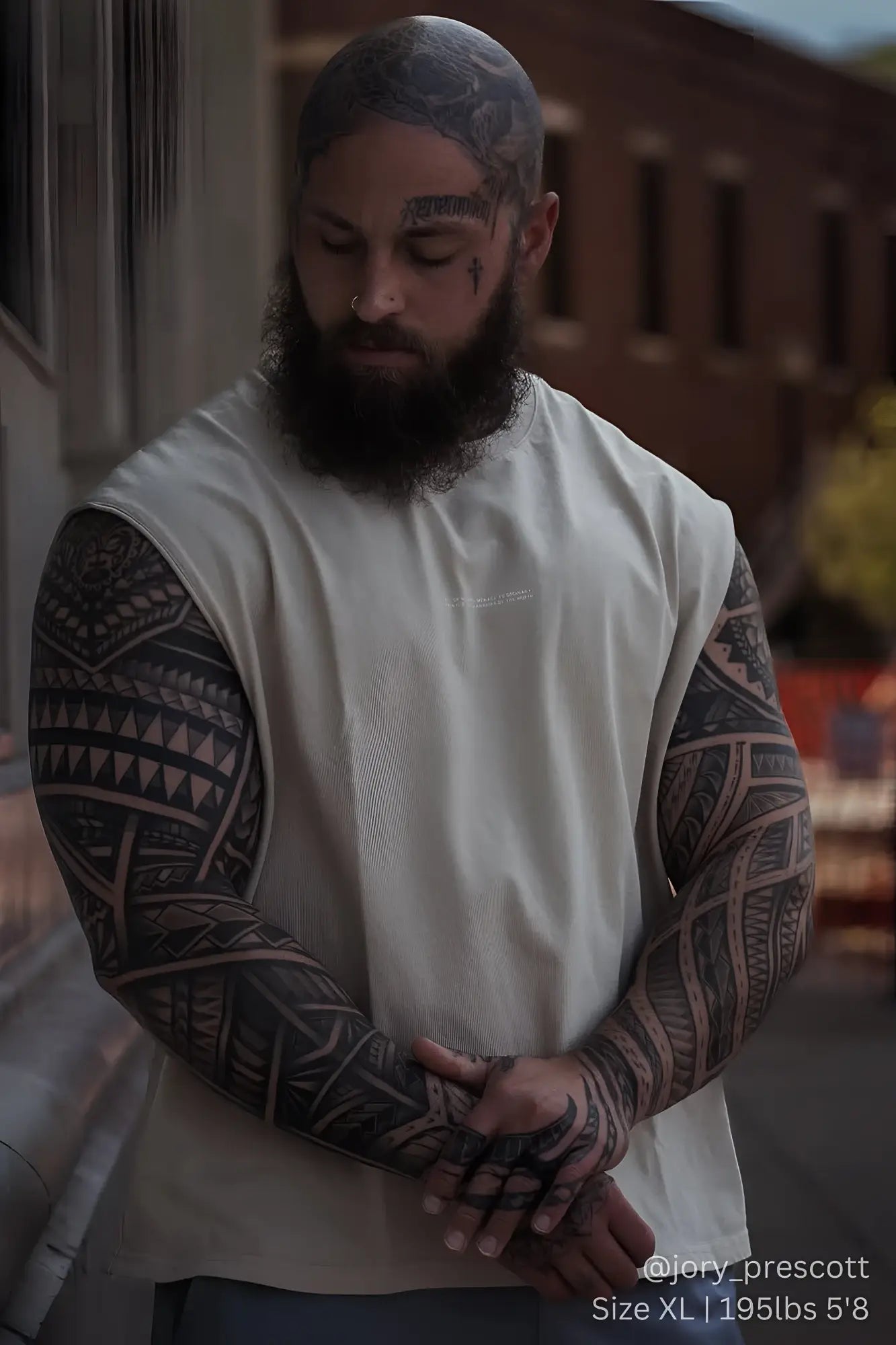 A man wearing a white sleeveless jed north sleeveless box cut tank top with black tribal tattoos on arms and head