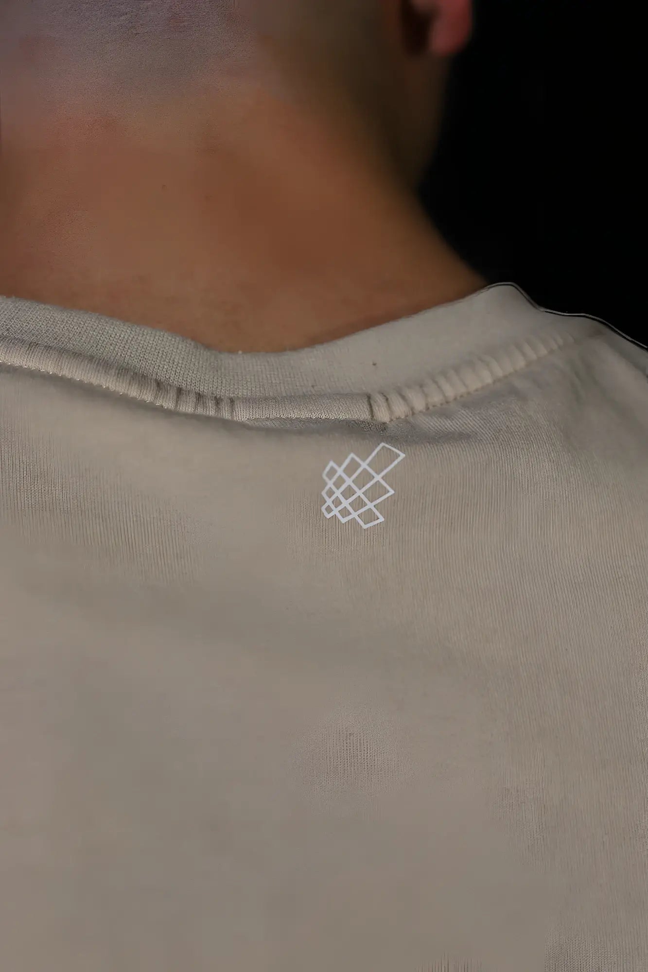 The back of a jed north sleeveless box cut tank top in light khaki showing a subtle white geometric logo embroidered on gray cotton
