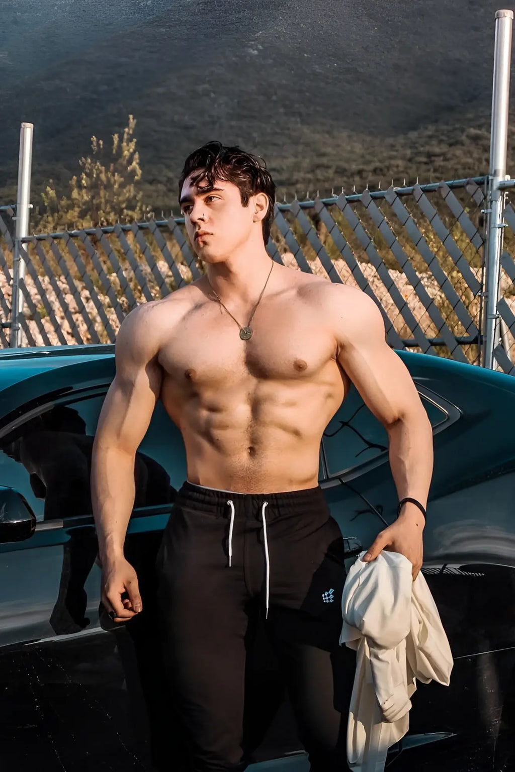 A muscular man wearing black spirit joggers with white drawstrings stands shirtless holding a light garment by a teal car