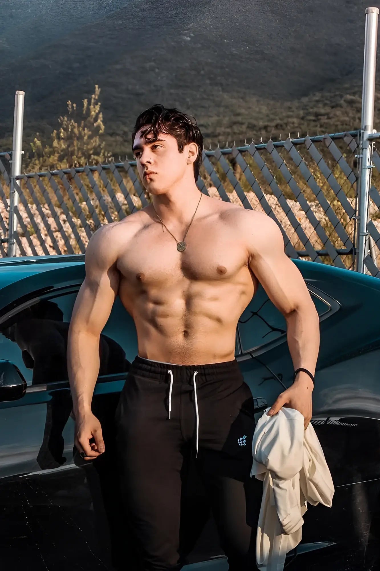 A muscular man wearing black spirit joggers with white drawstrings stands shirtless holding a light garment by a teal car