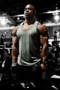 A muscular man wearing a light green Jed North Vital Raw-Hem Tank Top 2.0 holding dumbbells in each hand