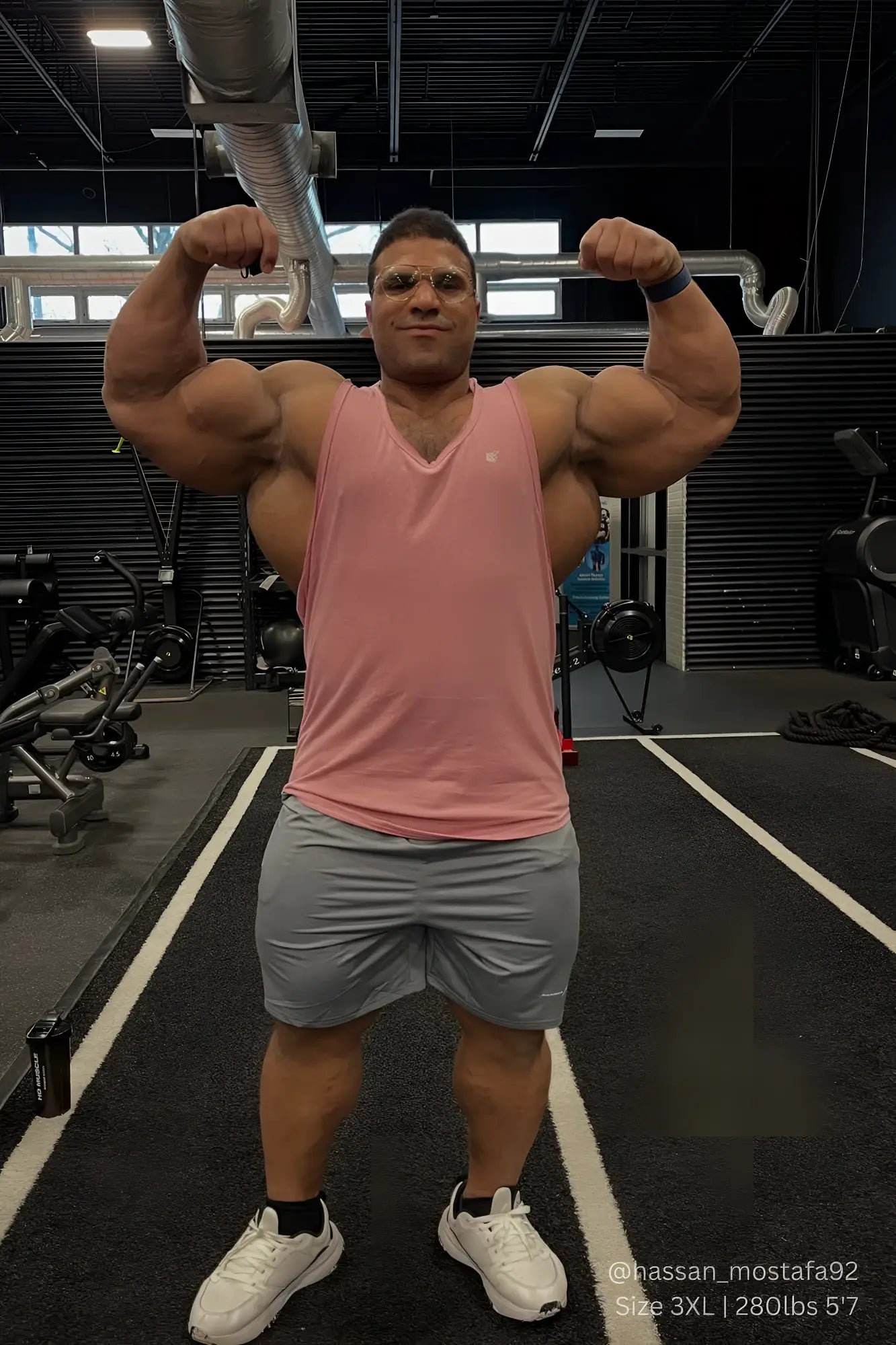 A muscular man flexing biceps in a jed north vital raw-hem tank top 2.0 salmon pink and gray gym shorts