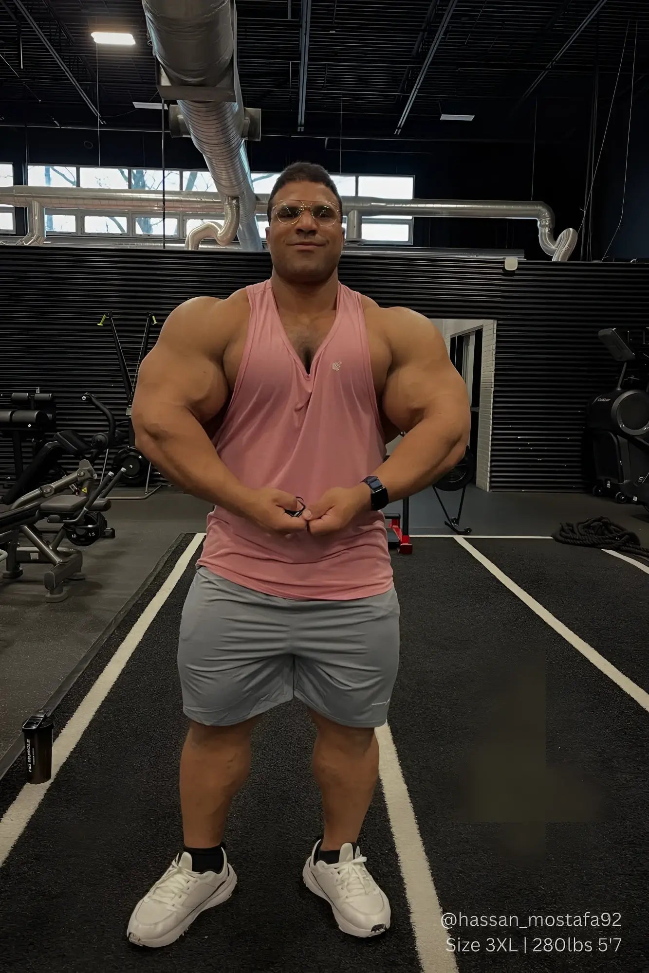 A muscular man wearing a salmon pink Jed North Vital Raw-Hem Tank Top 2.0 and gray shorts stands in a gym