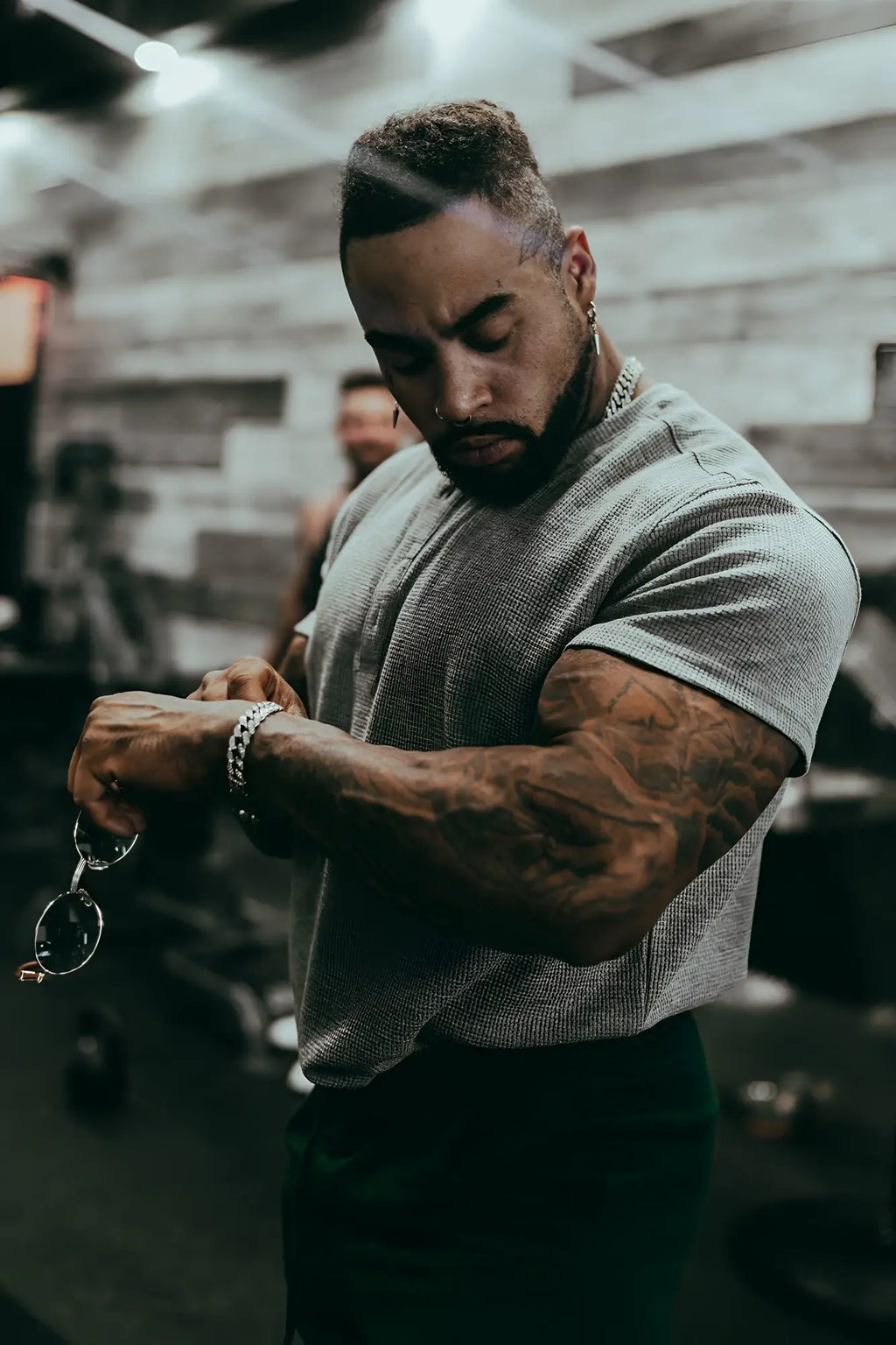 A man with tattoos wearing a light gray waffle henley t-shirt adjusting his watch and holding silver-framed glasses
