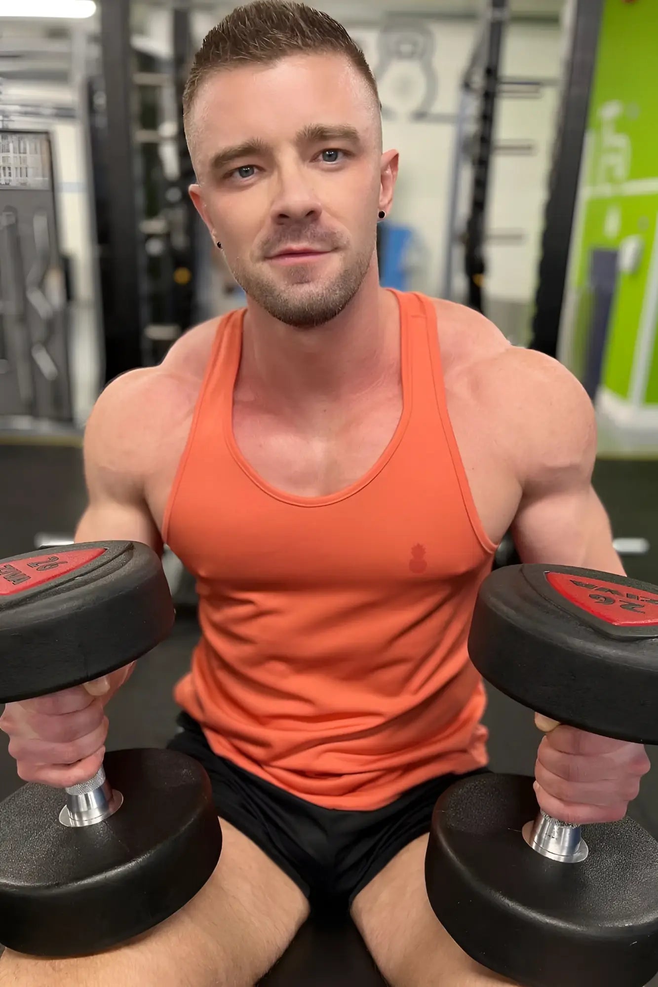 A man in a coral jj cool-dry gym stringer tank top holds black dumbbells with red weight plates