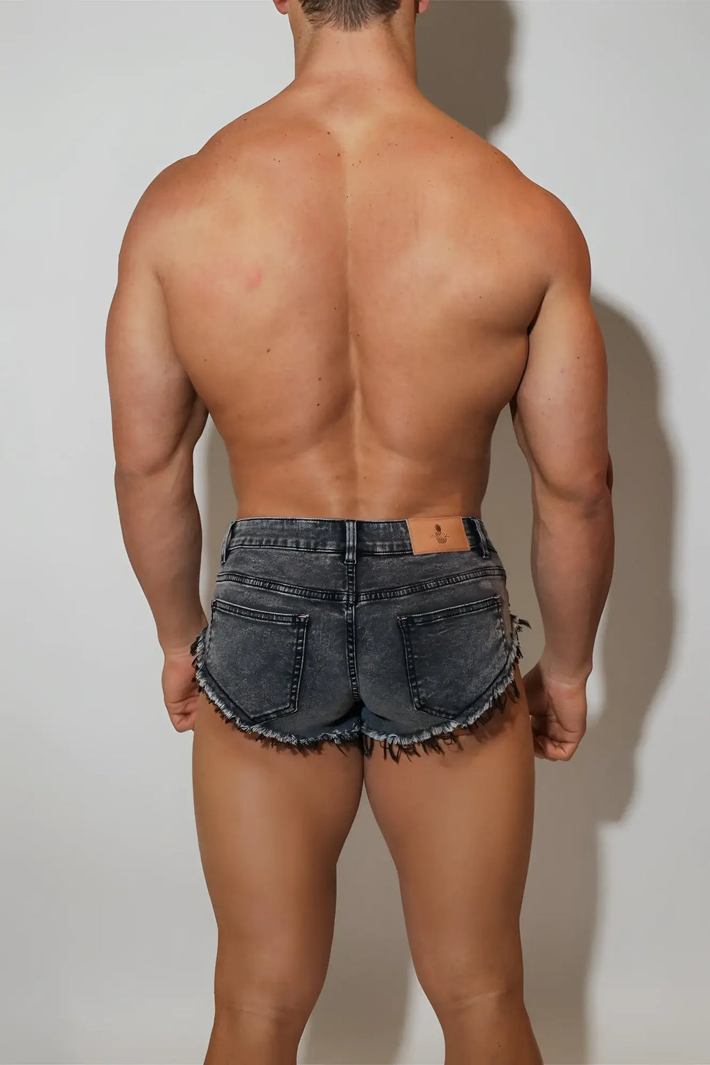 Acid washed distressed booty shorts in black dark-wash denim with frayed hem and brown leather patch at waistband back view