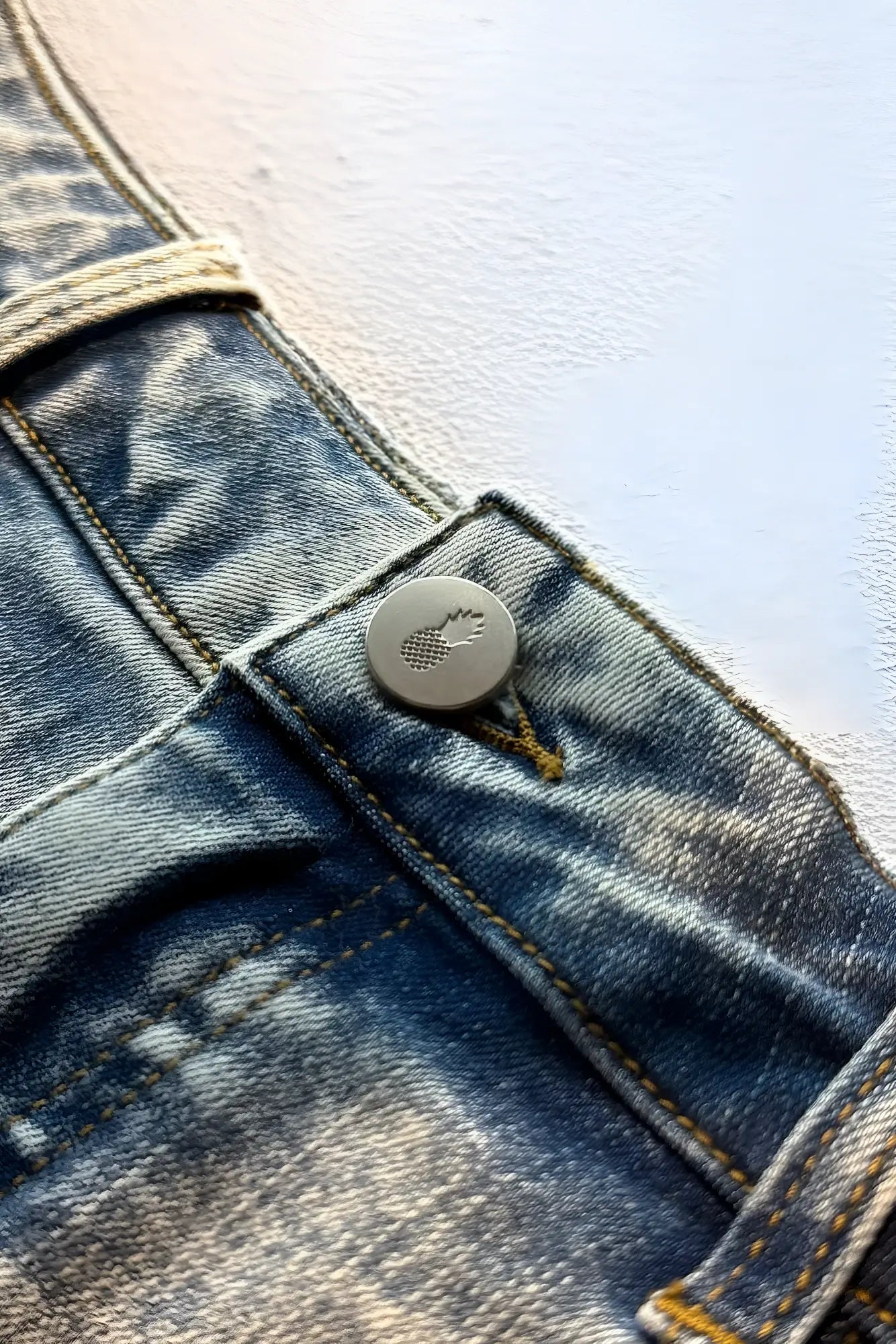 A round metallic button with an embossed logo on light blue acid washed distressed denim booty shorts back side