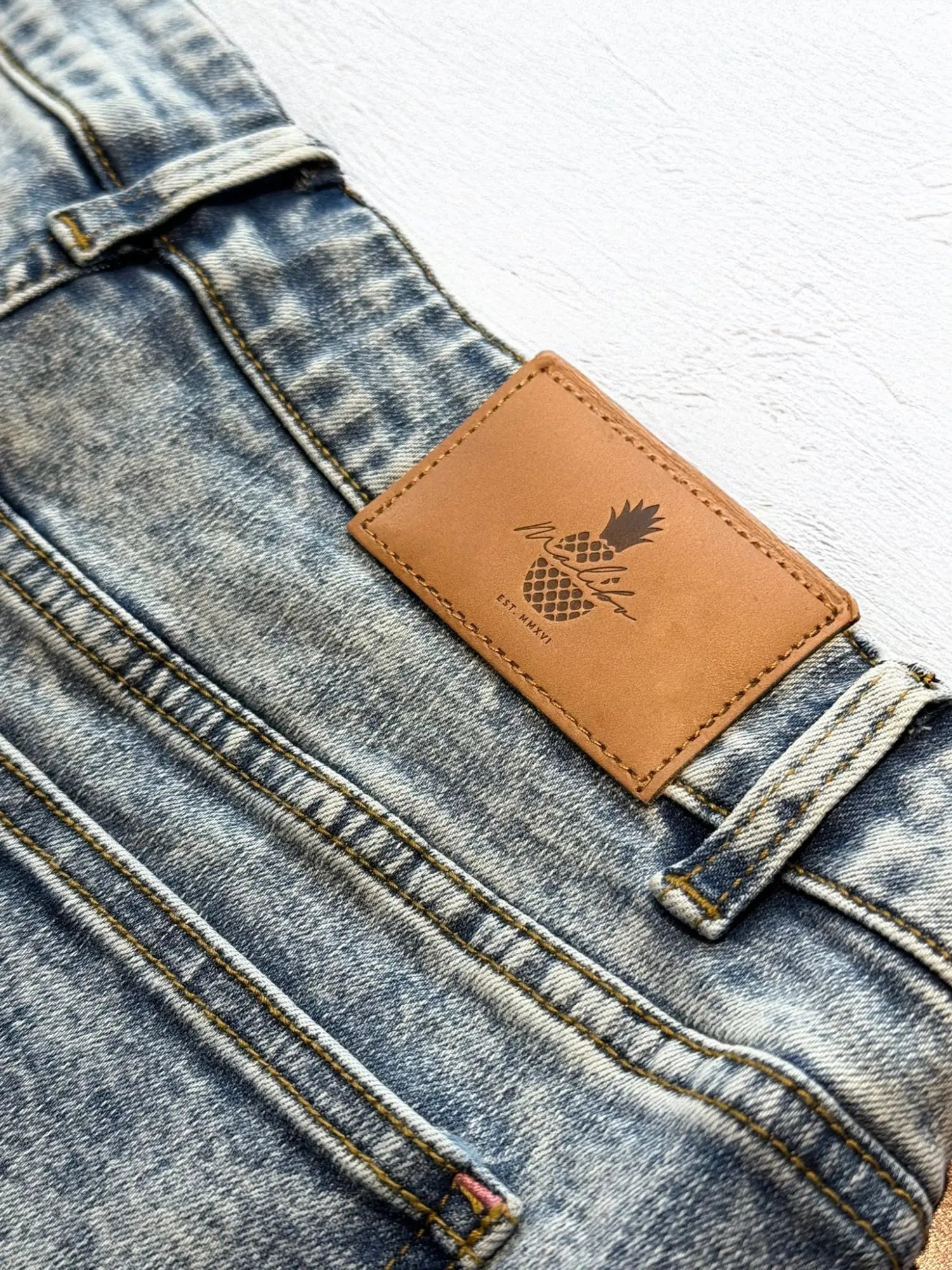 Light-wash acid washed distressed booty shorts with tan leather pineapple patch on the back waistband