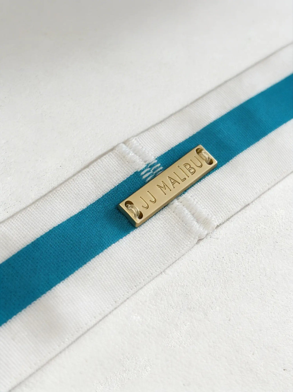 Blue on-display jockstrap with white fabric strap featuring bold teal stripes and gold-tone metal tag engraved jj malibu