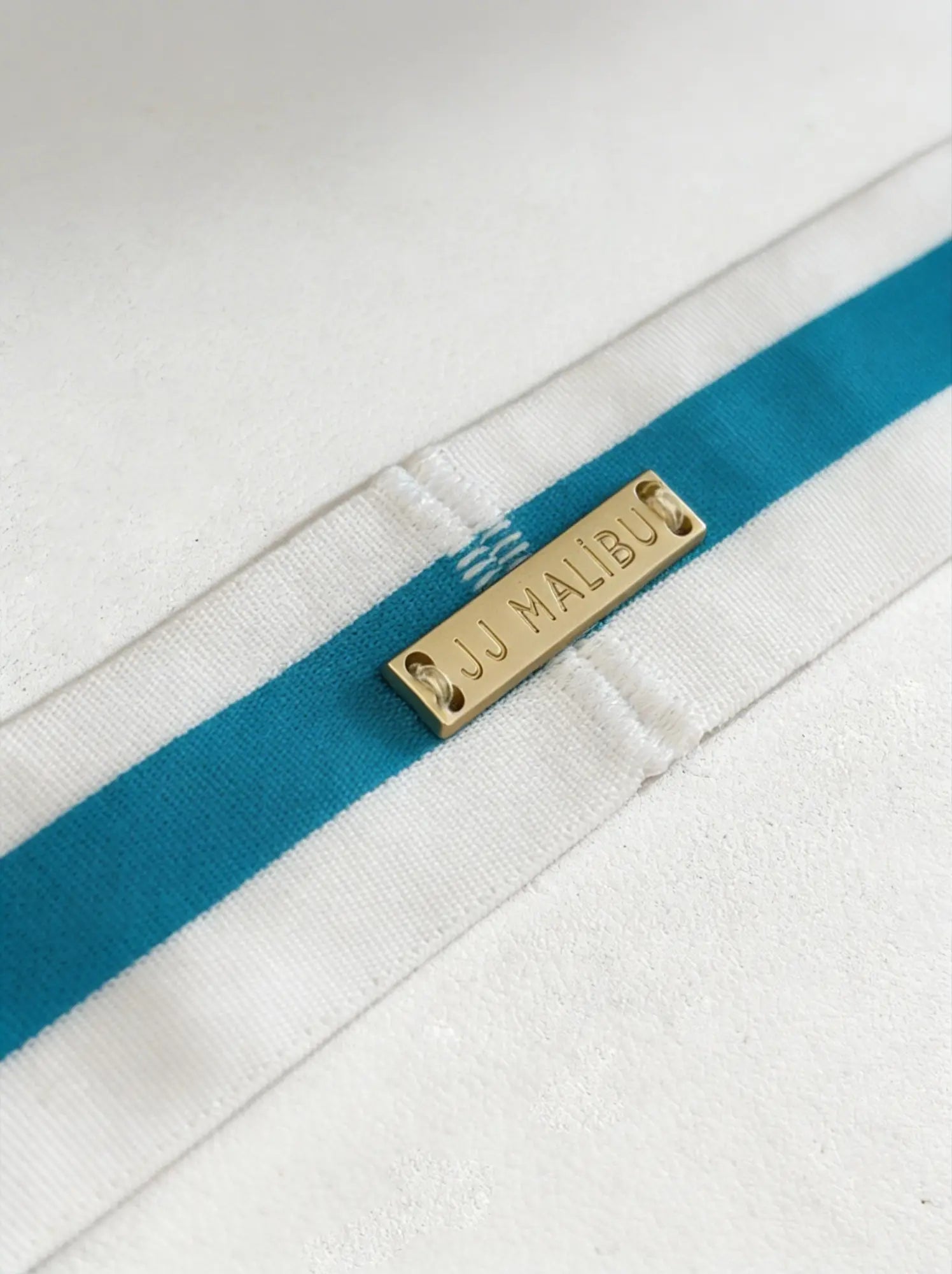 Blue on-display jockstrap with white fabric strap featuring bold teal stripes and gold-tone metal tag engraved jj malibu