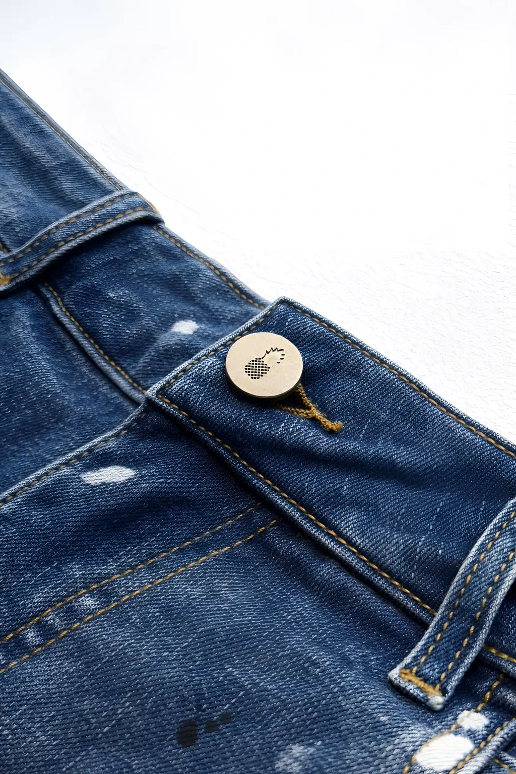 A circular metallic button with a textured foot logo sewn onto dark blue denim fabric of studio splash denim short shorts