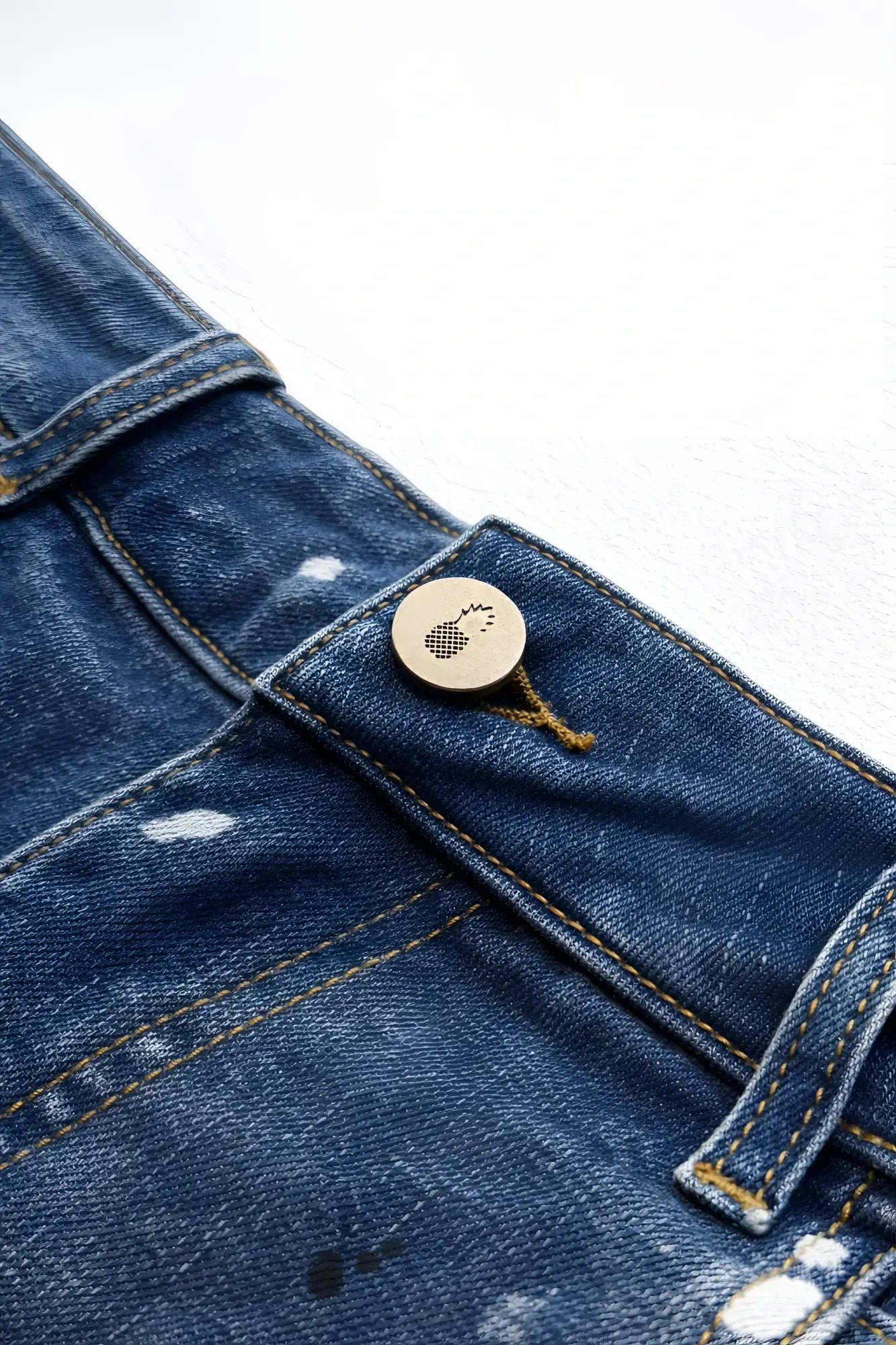A circular metallic button with a textured foot logo sewn onto dark blue denim fabric of studio splash denim short shorts