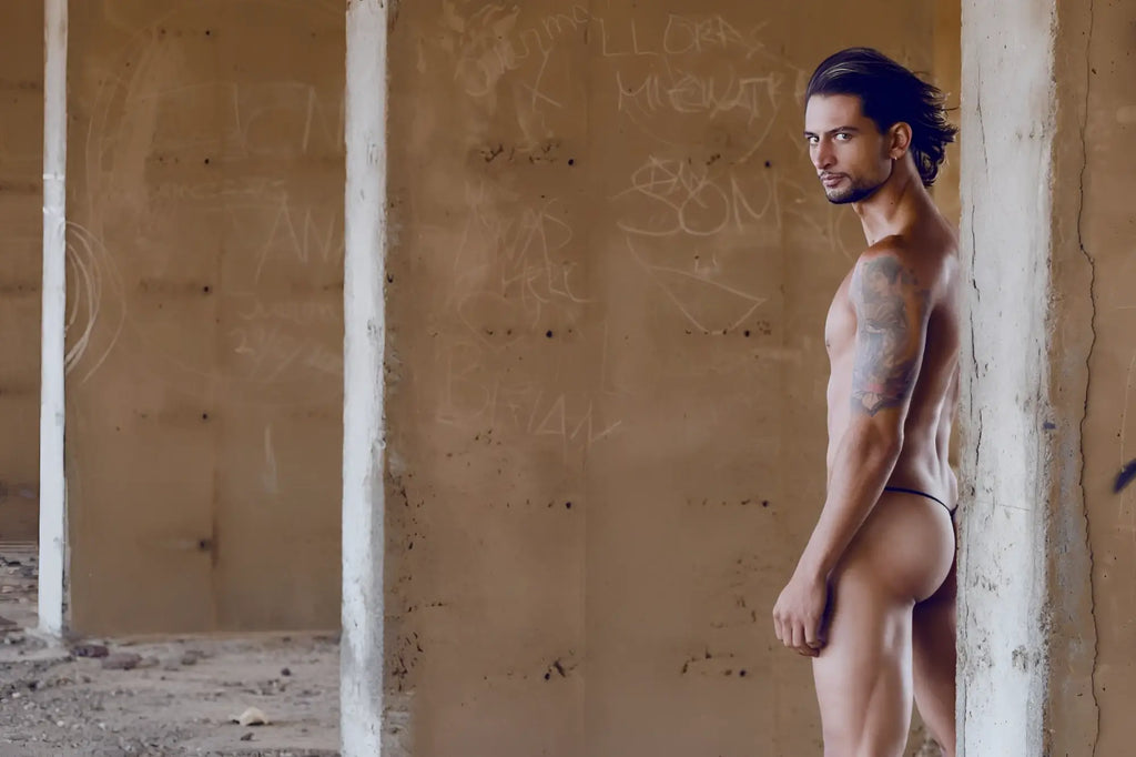 A muscular man with long dark hair and tattoos stands nude wearing a turquoise justin simon xsj02 classic g-string bulge