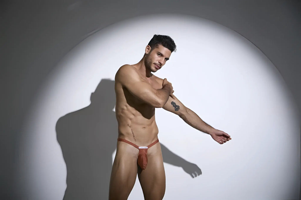 A man wearing a reddish brown thin strapped jockstrap with a small white label in justin simon xsjbu02 ochre color