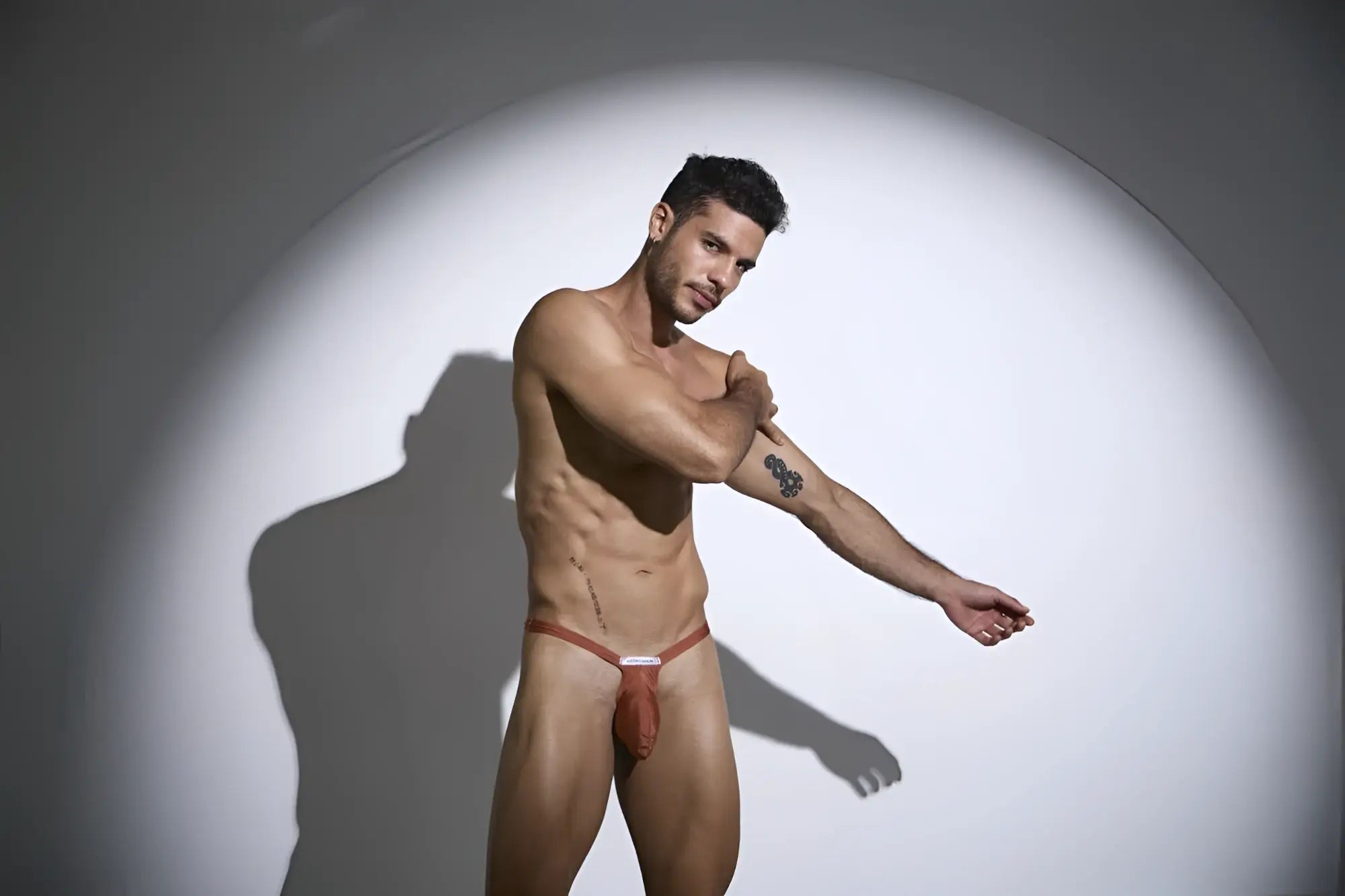 A man wearing a reddish brown thin strapped jockstrap with a small white label in justin simon xsjbu02 ochre color