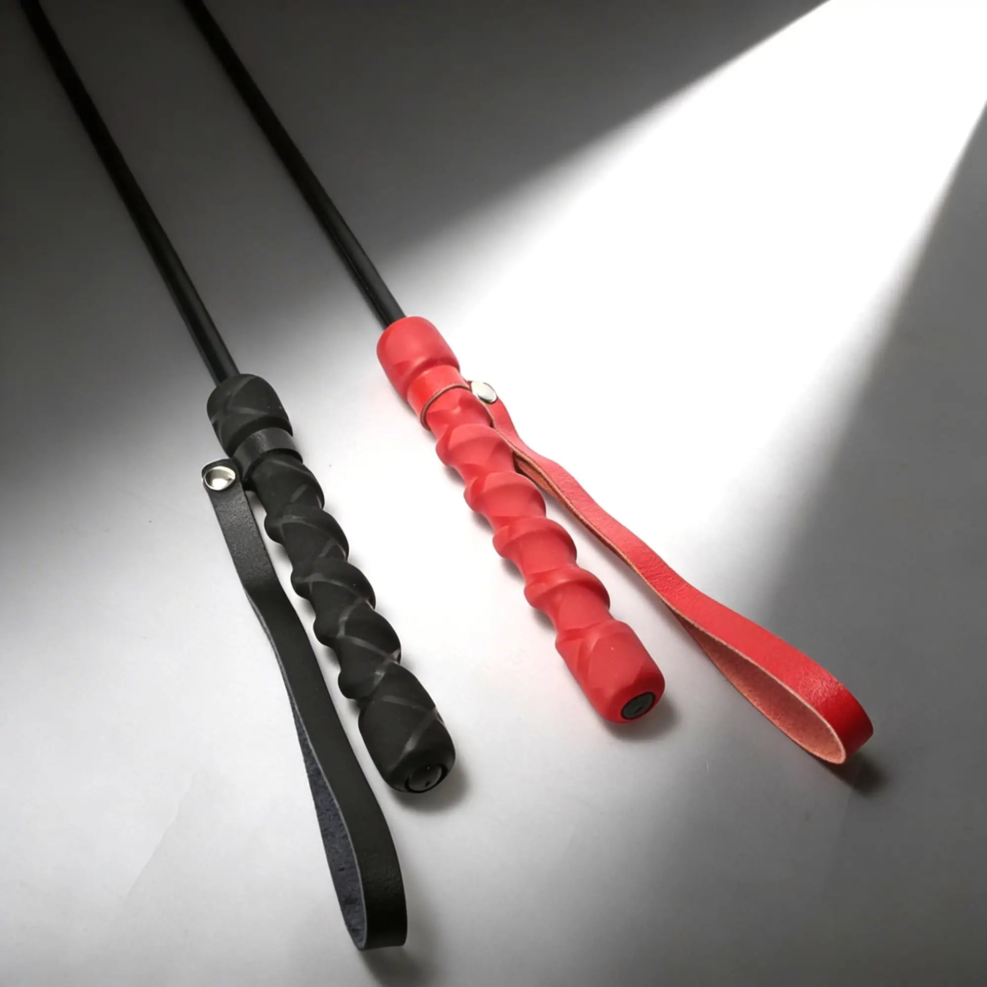 Two riding crops with textured, spiral-grip handles in black and red, each featuring an attached leather wrist loop.
