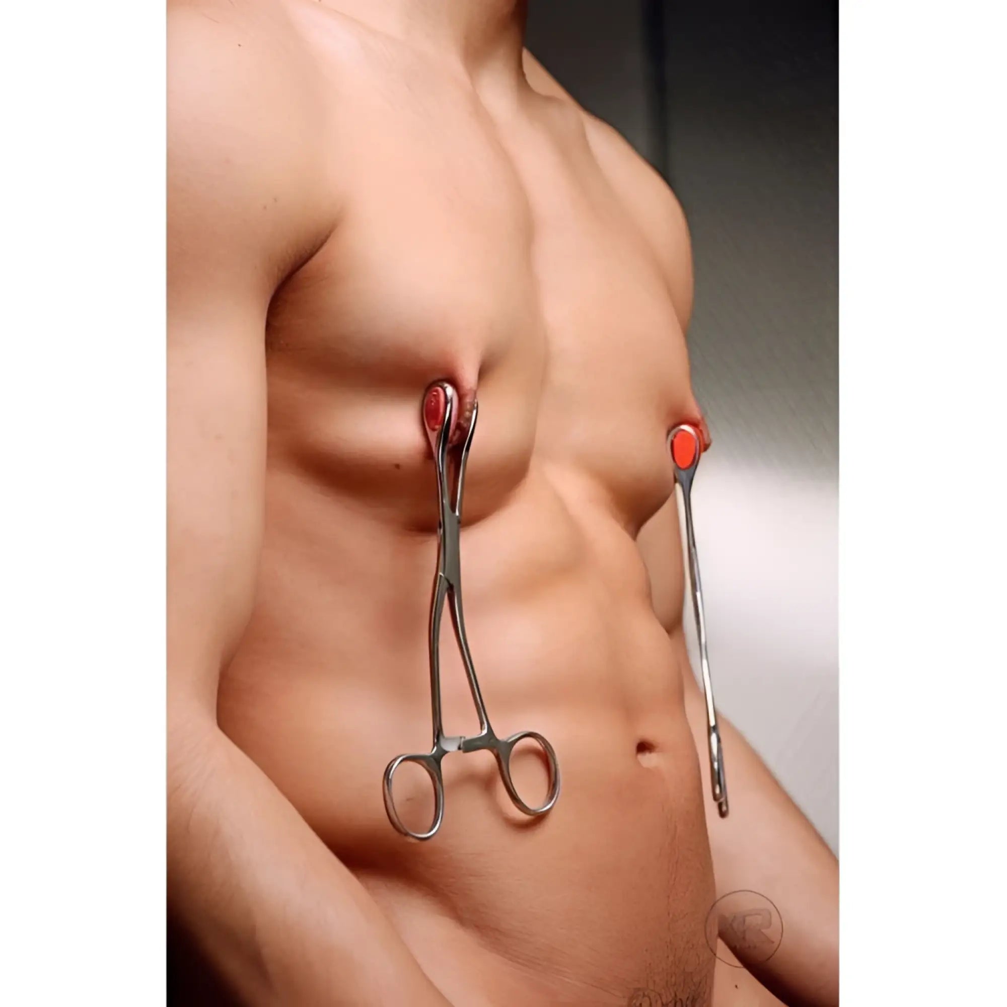 Kink Industries Young Forceps: A shirtless man wearing two polished metal medical-style nipple clamps with red pads.