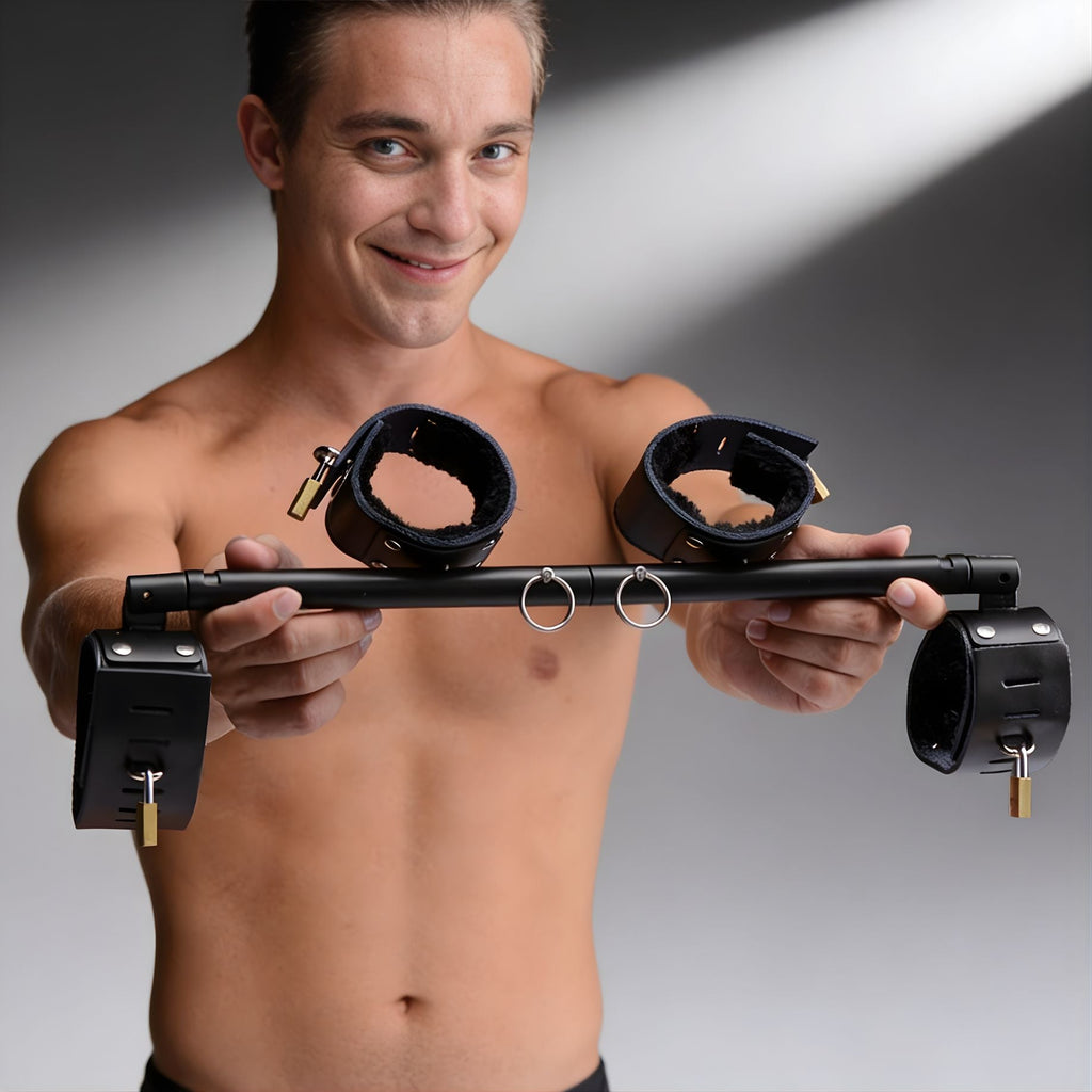 A shirtless man holds a black Strict Leather Locking Spreader Bar with four padded cuffs and brass padlocks.