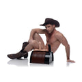 A man in a cowboy hat and boots sitting next to the LoveBotz Saddle, a brown and black faux-leather sex machine.