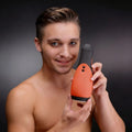 A shirtless man smiles while holding a black and orange LoveBotz Vibra Head Bionic Auto Stroker against a dark background.
