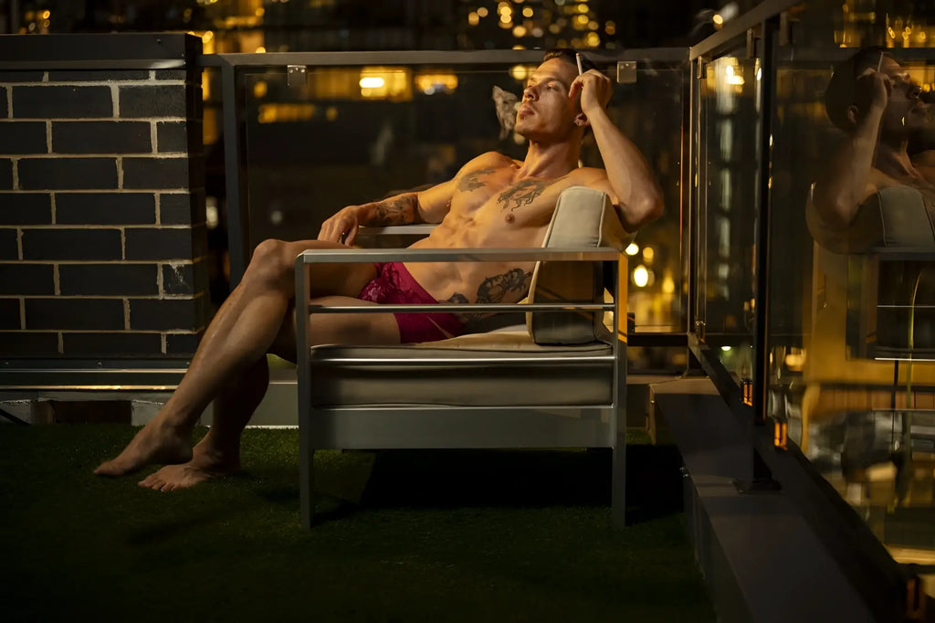 A man with tattoos lounges in a modern metal-framed chair wearing male power 126-289 lucifer cut out short in burgundy