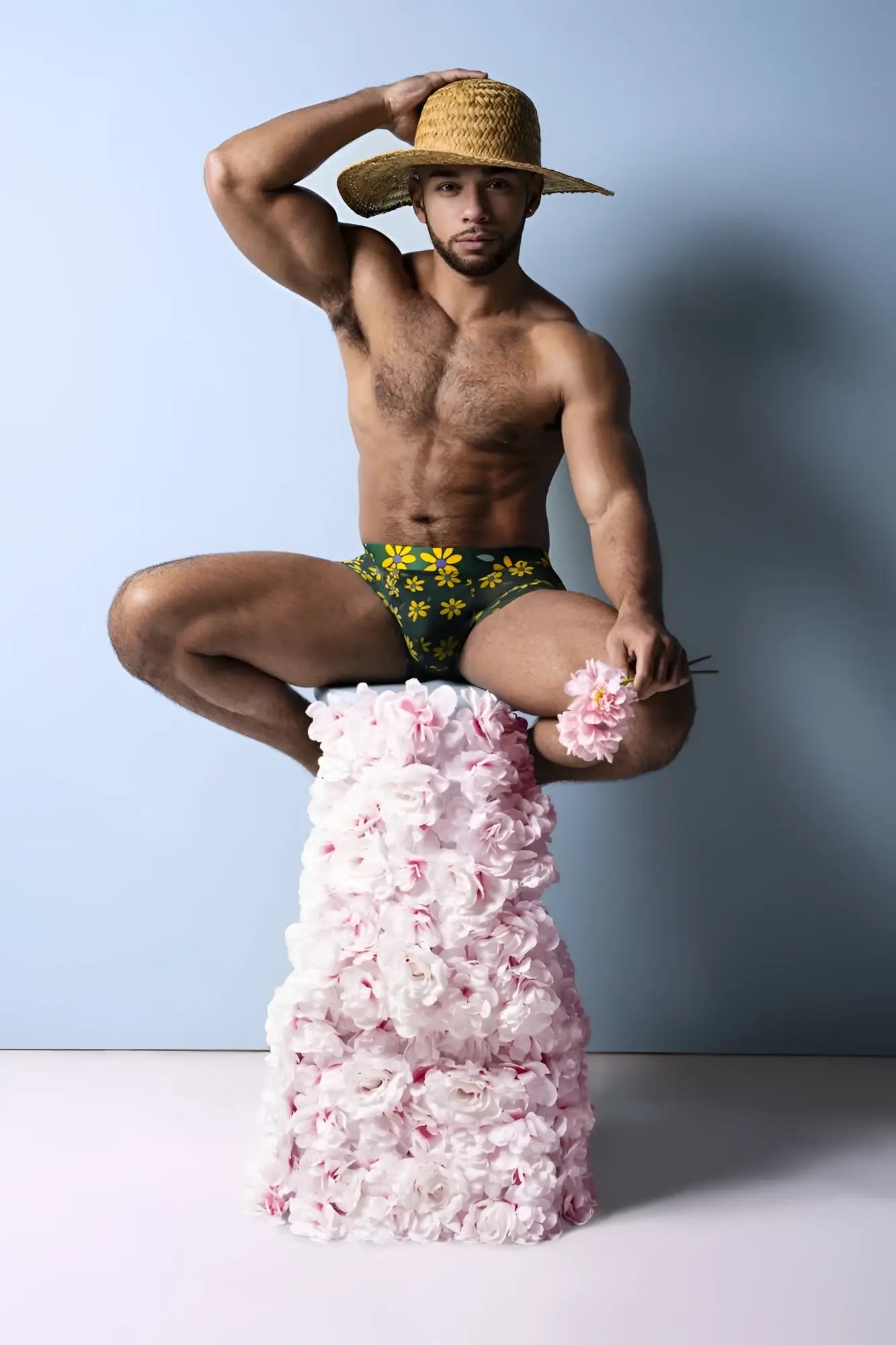 A man wearing a straw hat sits on a pedestal of pink roses wearing male power petal power pouch shorts in green daisy print