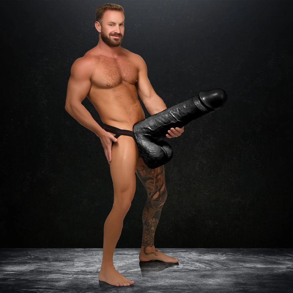 A muscular man holds a black, two-foot-tall Master Cock Moby super dildo in front of a dark background.