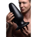 A man holds a smooth, black Master Cock Gigantor XXXL, a large, tapered silicone butt plug with a flared base.