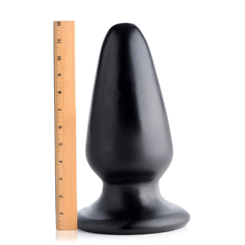 Master Cock Gigantor XXXL black tapered butt plug next to a 12-inch wooden ruler for scale on a white background.