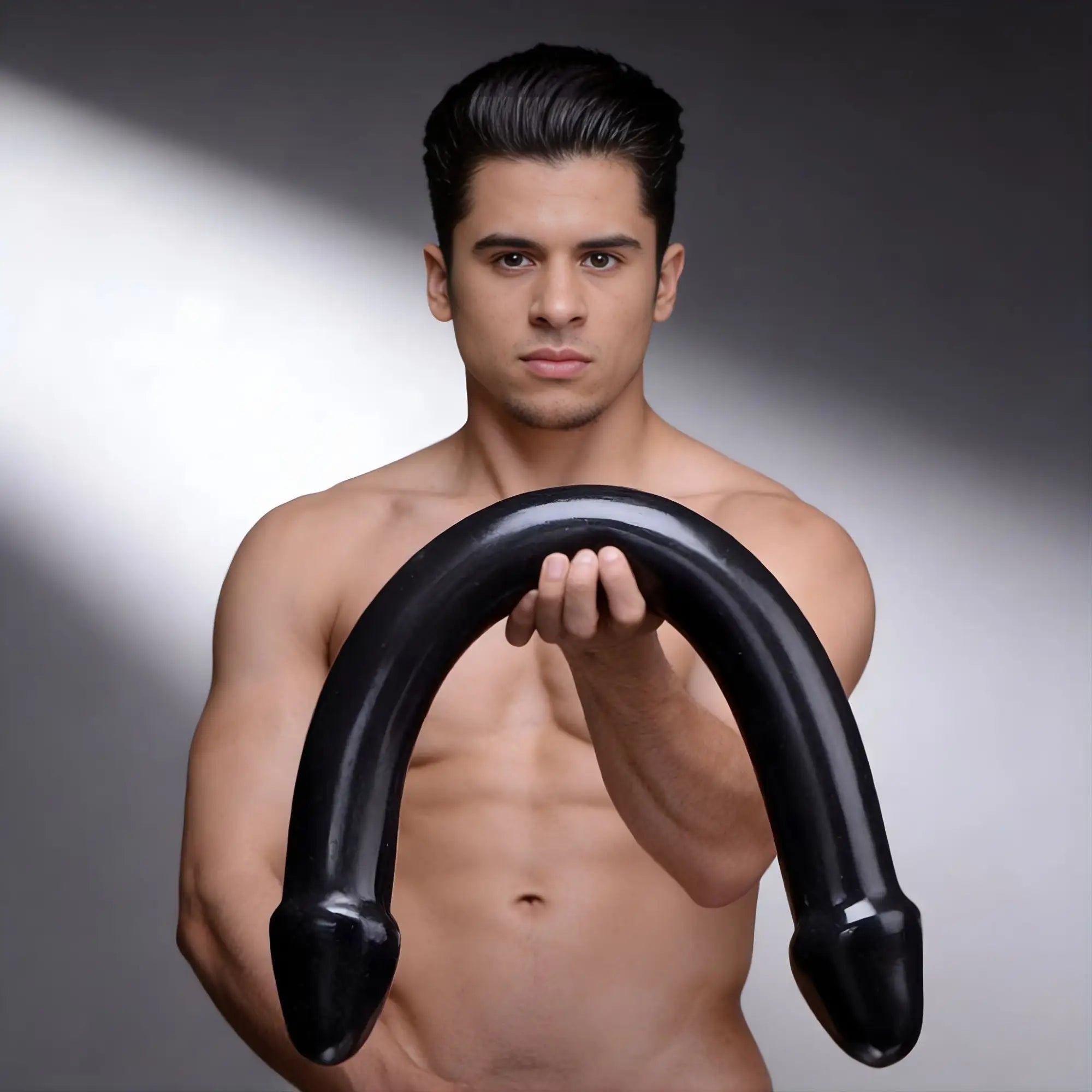 A shirtless man holds a black Master Cock The Python XL double-ended, U-shaped silicone dildo against a grey background.