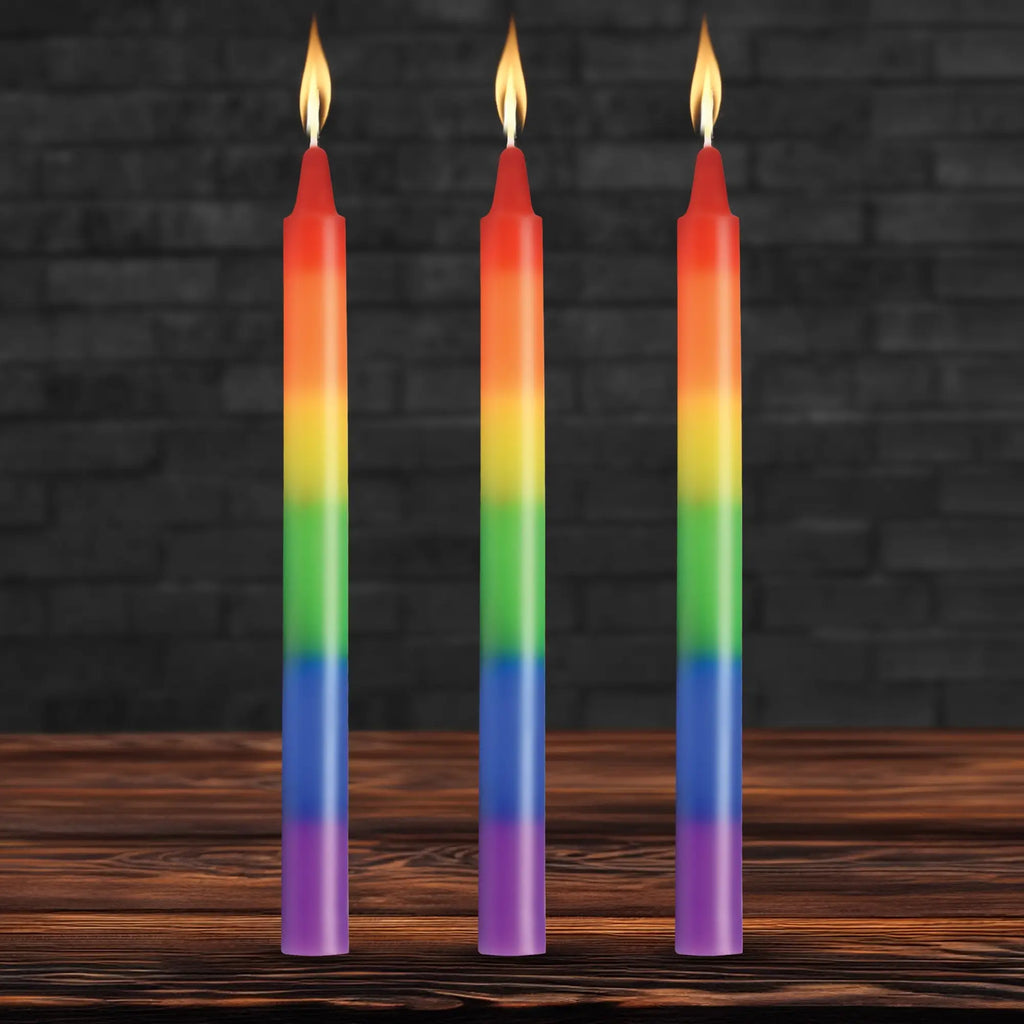 Three Master Series Pride taper candles in rainbow stripes, each with a lit flame, standing on a wooden surface.