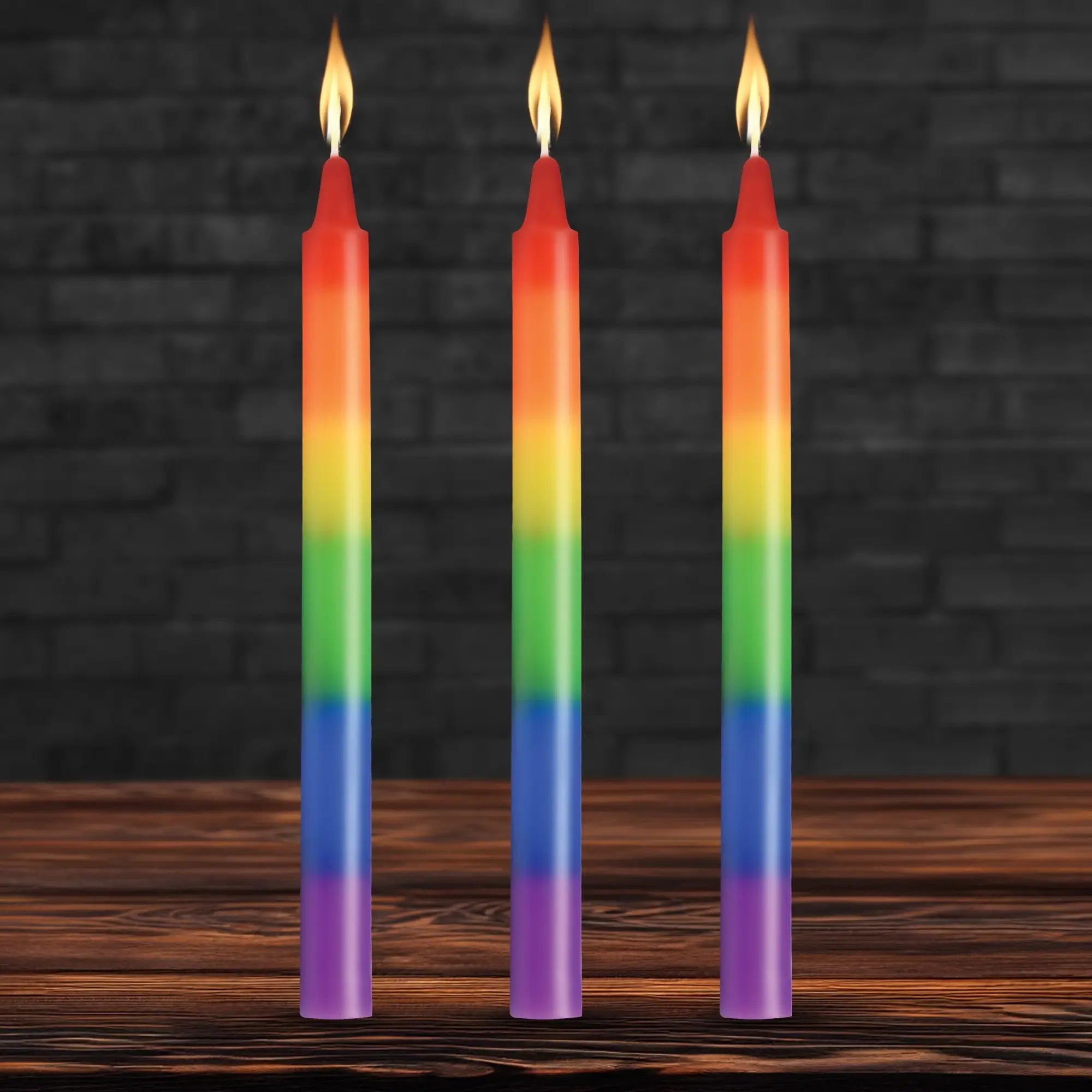 Three Master Series Pride taper candles in rainbow stripes, each with a lit flame, standing on a wooden surface.