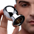 A hand holds a shiny, chrome-finished Master Series The Abyss Hollow Anal Dilator in front of a person's blurred face.