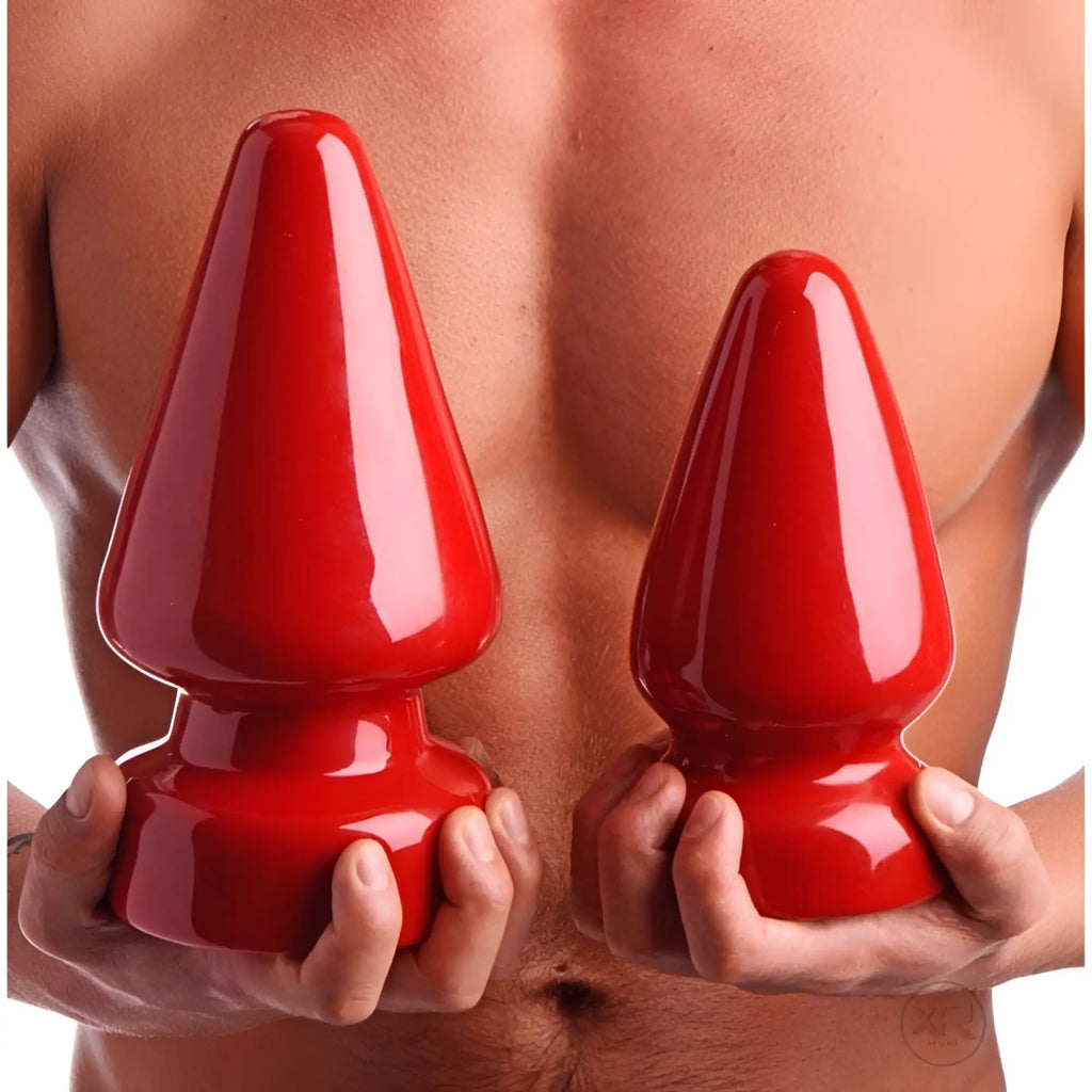 A person holding two large, glossy red Master Series The Anal Destructor butt plugs of different sizes.