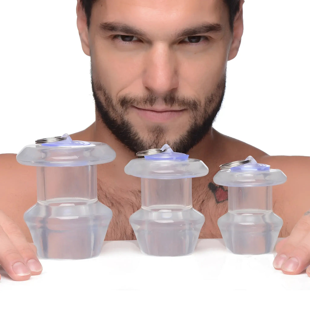 A man displays three clear, hollow Master Series Ass Bung anal dilators with blue plugs in varying sizes.