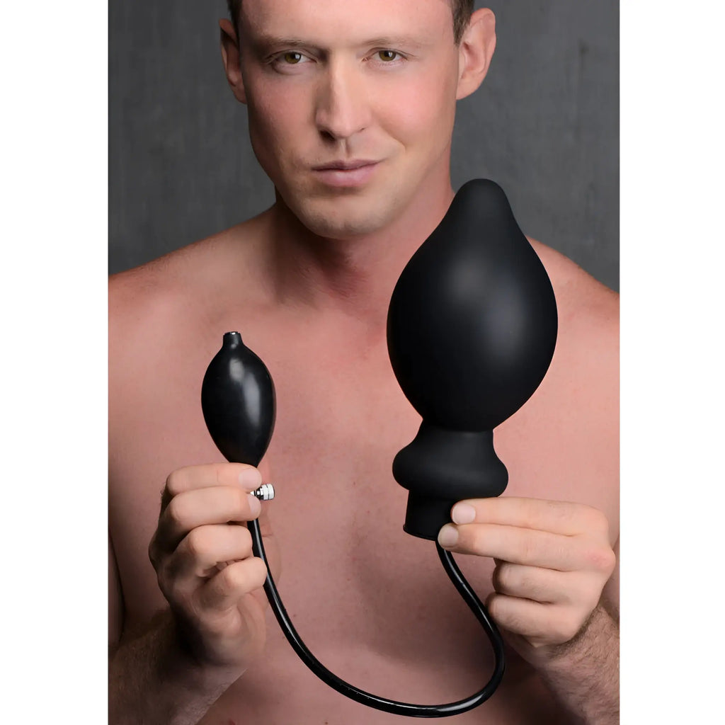 A man holds a large, matte black, inflatable silicone Master Series Ass-Pand anal plug with an attached hand pump.