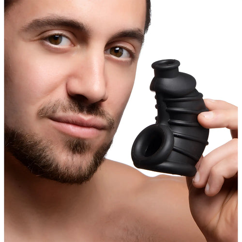 A man holds a black, ribbed Master Series The Chamber silicone chastity cage against a white background.