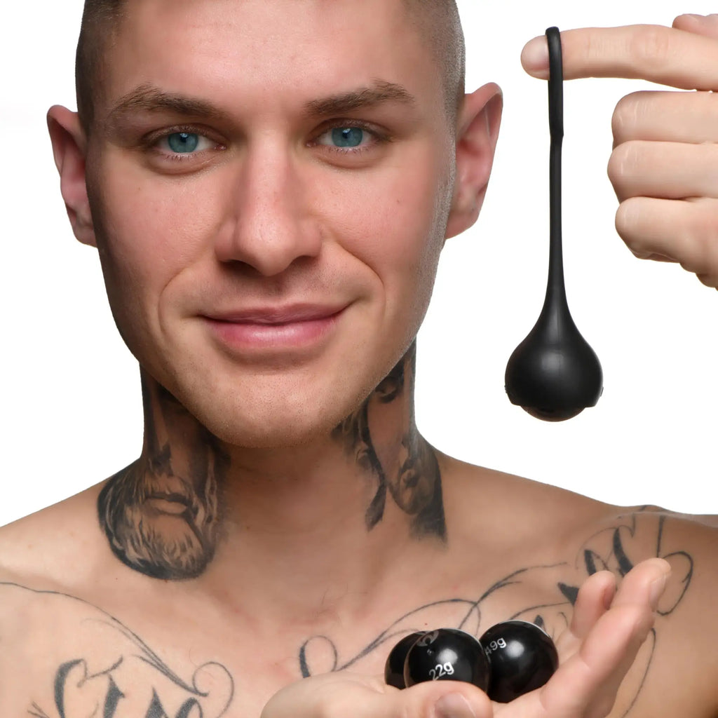 A tattooed man holding a black silicone Master Series Cock Dangler strap and matching spherical weights.