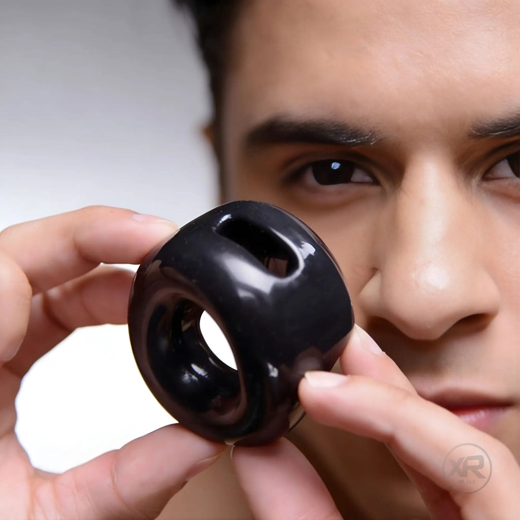 A man holds a smooth, glossy black Master Series Cock Holster cock and ball ring up to his face.