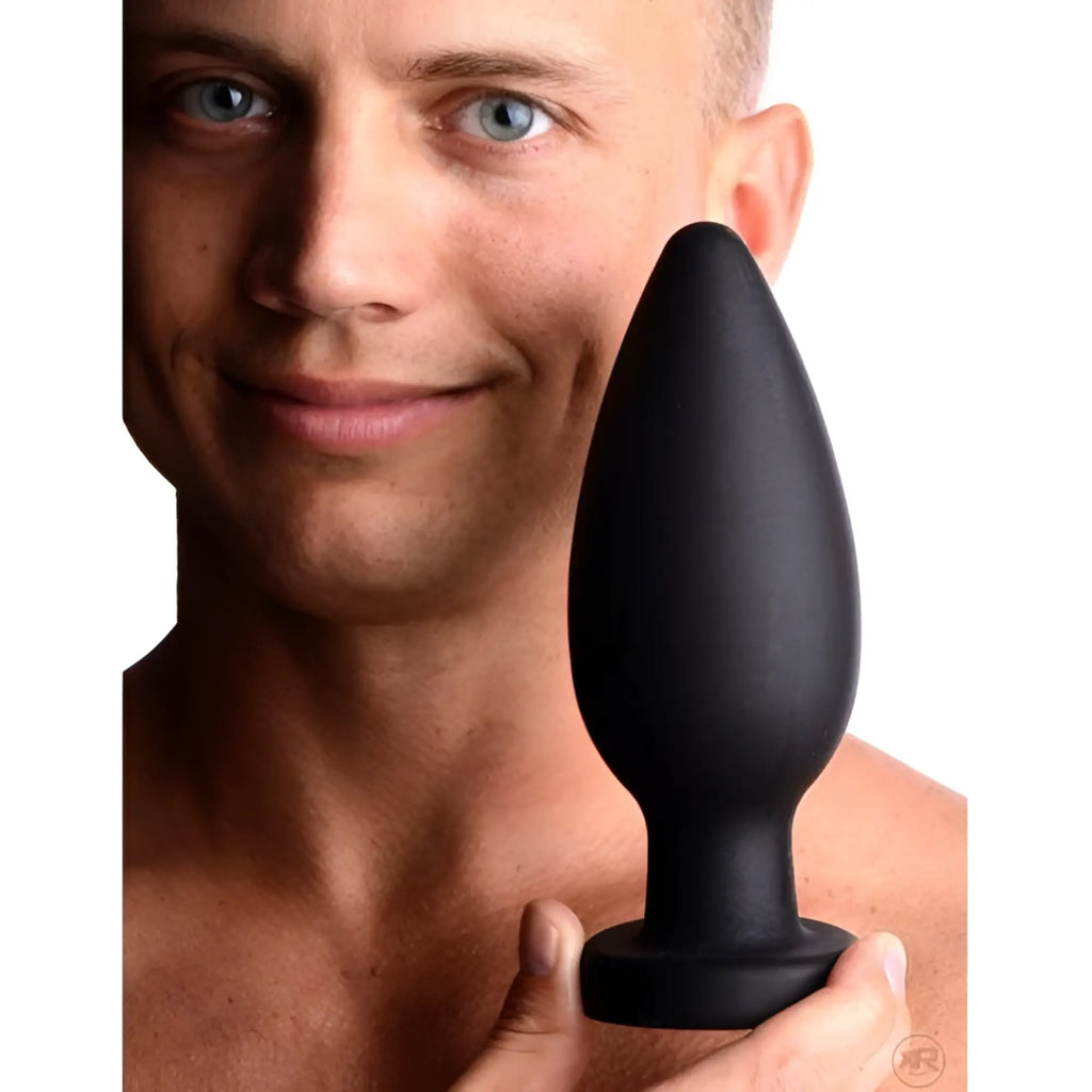 A man holds a smooth, black silicone Master Series Colossus XXL suction cup plug in front of his face.