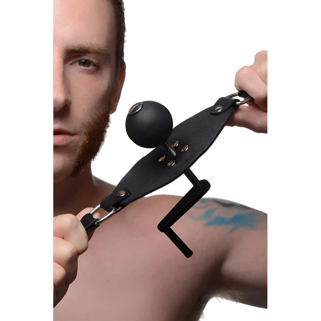 Master Series Crank Ball Gag with a black ball and adjustable leather strap held by a person.